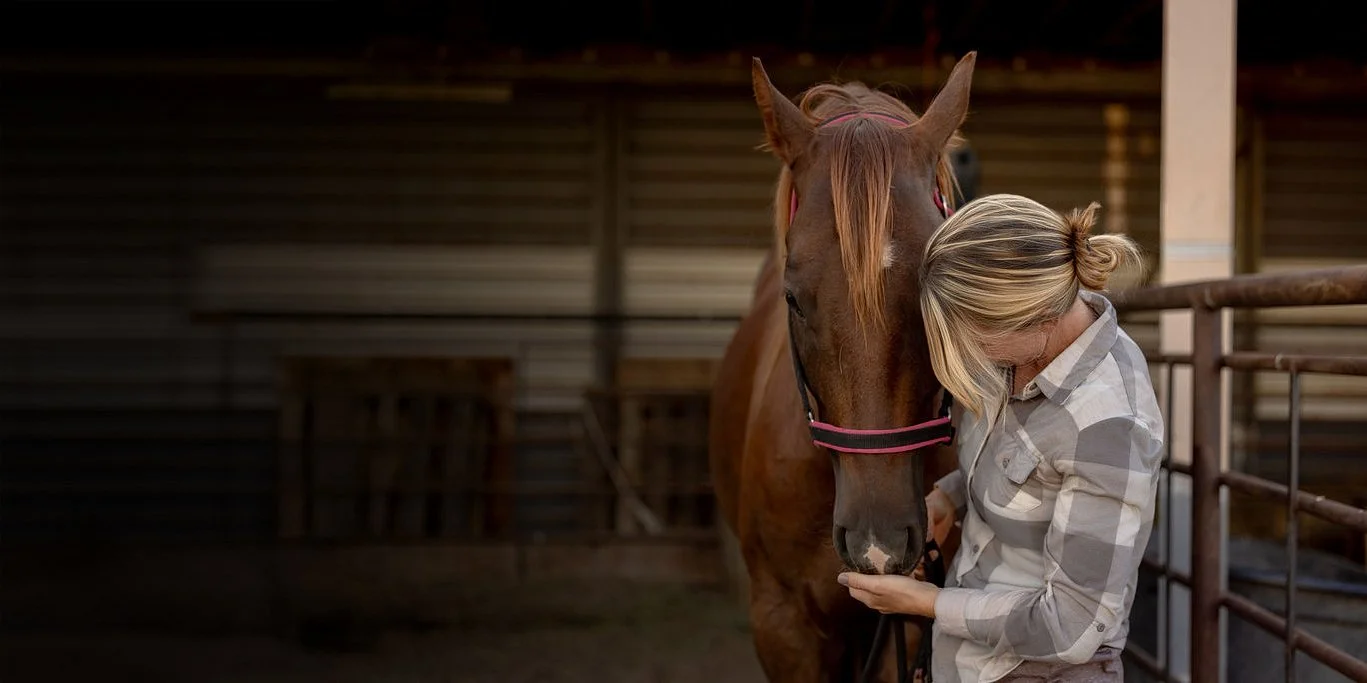Nicole resting her forehead against a horse in a quiet barn